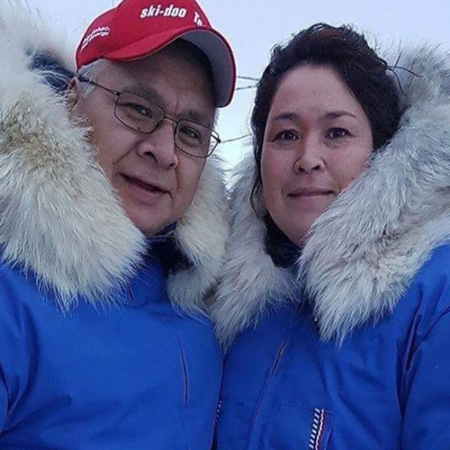 A man and a woman in blue fur-lined coats stand together in a snowy environment.